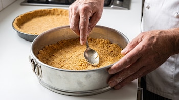 Manos de persona compactan masa de galleta en molde de tarta con cuchara metálica. Otra base de galleta visible al fondo sobre encimera.