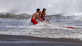 Dos jóvenes sin camiseta y una persona pequeña están en el mar con grandes olas, cerca de la orilla, junto a un flotador de rescate rojo