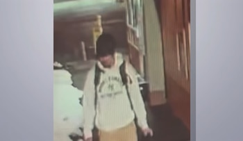 Hombre joven con un gorro oscuro, una sudadera con capucha de color claro con un logo, pantalones claros y una mochila oscura, caminando en lo que parece un pasillo o túnel de baja resolución