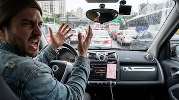 Hombre con gorro dentro de un auto, levanta las manos frustrado por el tráfico visto a través del parabrisas con gotas de lluvia y un teléfono con mapa.