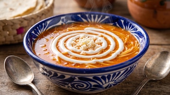 Tazón de cerámica azul con sopa de fideo, crema en espiral, queso y dos cucharas sobre una mesa de madera.