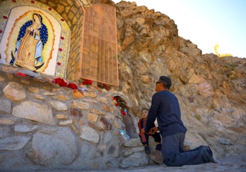 Hombre arrodillado con niño pequeño frente a un santuario de la Virgen de Guadalupe en una ladera rocosa. En la cima se ve una estatua amarilla