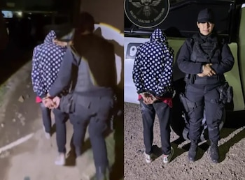 Una persona esposada con capucha de lunares permanece junto a una oficial de policía frente a un vehículo oficial durante la noche