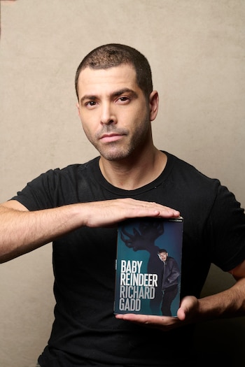 Un hombre con cabello corto y barba ligera, vestido con una camiseta negra, sostiene un libro titulado "Baby Reindeer" frente a un fondo beige
