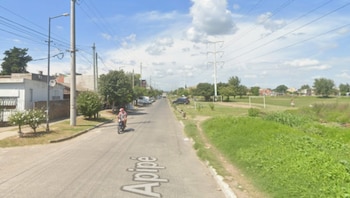 Calle Apipé con una persona en moto, casas a la izquierda, árboles, postes de luz y un campo de pasto con un arco de fútbol a la derecha bajo cielo nublado