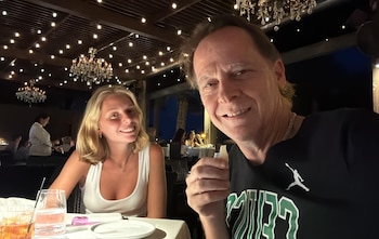 Hombre y mujer sonríen en primer plano. Al fondo se observa un restaurante con mesas, personas, luces colgantes y arañas de cristal