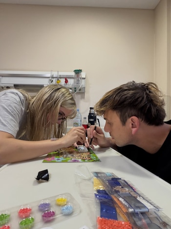 Hombre y mujer sentados frente a una mesa blanca, realizando una manualidad con pequeñas piezas de colores. Hay botellas, un termo y equipo médico en la pared