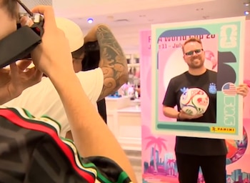 Persona fotografiando a un hombre sonriente con gafas de sol, sosteniendo un balón de fútbol y un marco de fotos de la Copa Mundial 2026 en una tienda