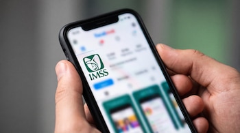 Mano sosteniendo un teléfono inteligente que muestra el logotipo verde del IMSS en la pantalla con una interfaz de redes sociales desenfocada y fondo gris.