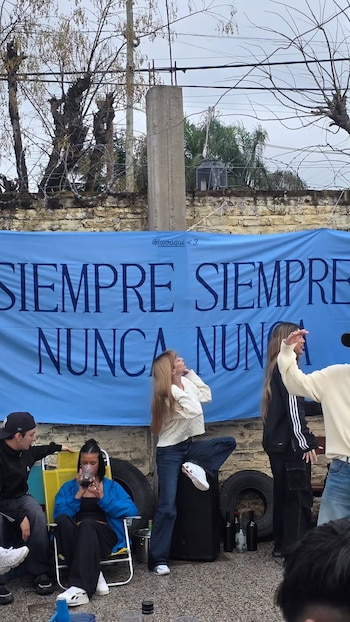 Una mujer de pie, otras personas sentadas, un gran cartel azul con texto, un muro de piedra, árboles sin hojas y alambre de púas