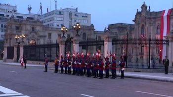 Grupo de soldados con uniformes rojos y azules, con banderas peruanas, formados frente al Palacio de Gobierno y su reja. Edificios y farolas visibles