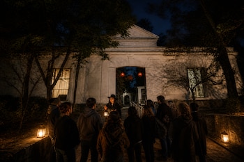 Personas reunidas de noche frente a la fachada blanca de un edificio antiguo, mientras una mujer con sombrero sostiene un farol encendido.