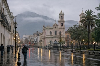 Calle adoquinada y mojada con personas que llevan paraguas, edificios de estilo colonial, faroles encendidos y una montaña con niebla al fondo.