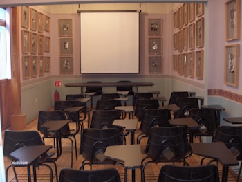 Interior de un aula con una pantalla de proyección blanca, una mesa larga al frente, filas de sillas negras con escritorios integrados y cuadros en las paredes