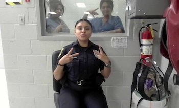 Una mujer con uniforme policial posa sentada haciendo gestos con las manos, mientras dos personas tras una ventana de cristal realizan señas