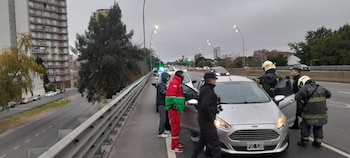 Vehículo Ford plateado en una autopista con personal de emergencia, incluyendo bomberos y un agente, y otros vehículos y edificios al fondo