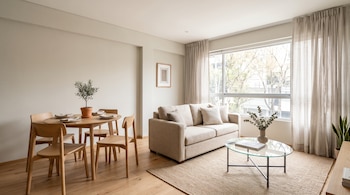 Comedor con mesa redonda de madera y sillas junto a una sala con sofá beige, mesa de centro de vidrio, alfombra y ventana amplia con cortinas.