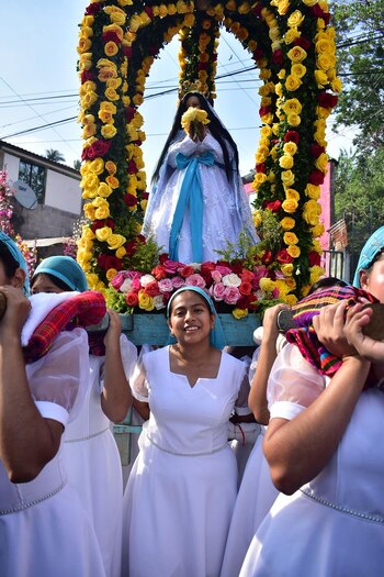 Estatua de la Virgen María en andas florales amarillas y rojas, portada por jóvenes sonrientes con vestidos blancos y pañuelos azules, bajo cielo azul