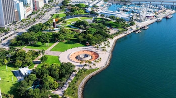Vista aérea de un gran parque verde junto al agua, con palmeras, edificios altos a la izquierda, y un muelle con barcos y una noria a la derecha