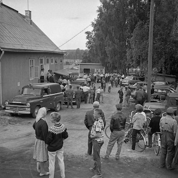 Imagen en blanco y negro de una multitud y varios coches, incluyendo un vehículo policial, reunidos en un camino de tierra frente a un edificio de madera con árboles