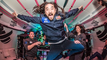 Steve Aoki flota con los brazos abiertos dentro de un avión, flanqueado por dos instructores con trajes de vuelo y luces rojas