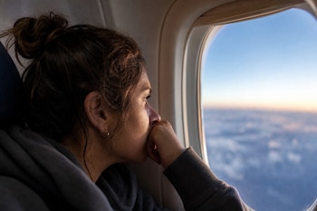 Perfil de una mujer con el rostro apoyado en su mano mientras mira por la ventanilla de un avión hacia las nubes.