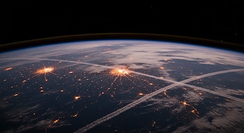 Vista aérea nocturna de la Tierra desde el espacio, mostrando grandes áreas urbanas con luces brillantes y estelas blancas de aviones cruzando la atmósfera terrestre.