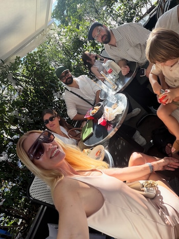 Una mujer rubia sonríe en primer plano con gafas de sol. Detrás, tres adultos y un niño con una manzana y cuchara en una mesa exterior con vegetación y toldo