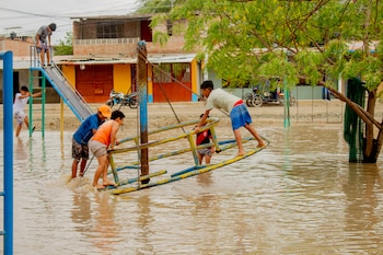 Varios niños permanecen sobre juegos metálicos en una zona recreativa cubierta de agua turbia, frente a casas y árboles
