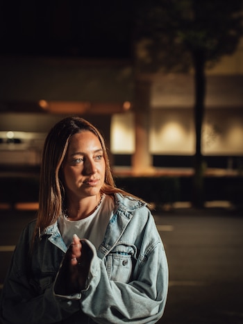 Mujer de cabello largo castaño y ojos claros con chaqueta de jean y camiseta blanca, posa de noche en un exterior con un árbol y luces de fondo