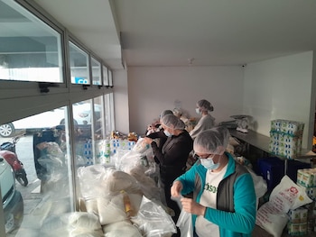 Varias personas con mascarillas y redecillas empaquetan productos en bolsas transparentes, rodeadas de grandes pilas de suministros dentro de un almacén