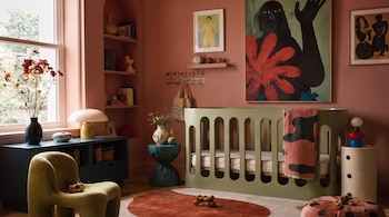 Dormitorio con cuna verde, alfombra roja y beige, sillón verde, estantes empotrados, cuadro con figura negra y flor roja, lámparas y varios objetos decorativos.