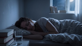 Mujer recostada en cama, durmiendo. Cabello oscuro y mojado. Vaso de agua en mesita de noche junto a pila de libros. Ventana con luna llena.