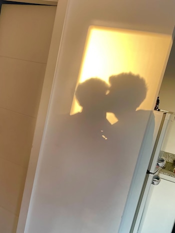 Siluetas de dos cabezas besándose proyectadas sobre un panel rectangular luminoso de color amarillo cálido, con una nevera blanca y pared clara a la derecha