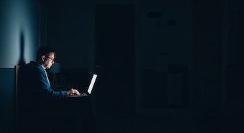 Vista lateral de una persona con gafas sentada en una habitación oscura, iluminada por la pantalla de una computadora portátil y una lámpara de mesa tenue.