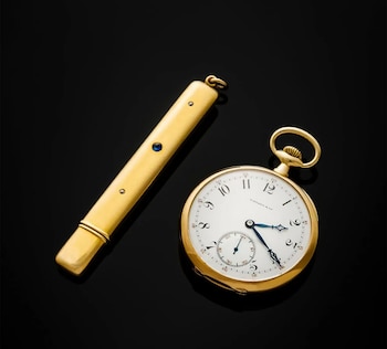 Primer plano de un reloj de bolsillo dorado con esfera blanca y manecillas azules junto a un objeto alargado dorado con gemas azules y claras sobre fondo negro