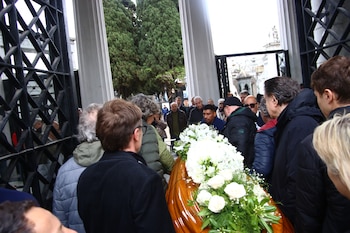 Varias personas rodean un féretro de madera cubierto de flores blancas frente a un pórtico con columnas en un cementerio