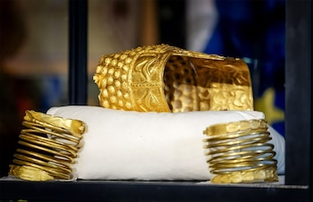 Un casco de oro elaborado con formas geométricas y dos brazaletes espirales de oro en un cojín blanco, sobre un fondo oscuro