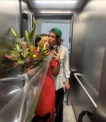 Reflejo en un ascensor de una mujer con vestido rojo de puntos blancos sosteniendo un ramo de flores, mientras un hombre con gorra verde la besa en la cabeza