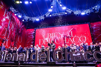 Escenario con un grupo de personas, pantalla roja con texto, letras grandes que forman Bienal del Chaco, y confeti cayendo sobre los presentes