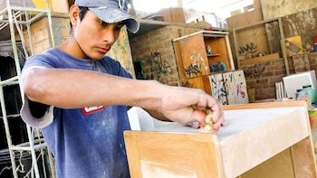Un joven con gorra y camiseta azul lija un mueble de madera en un taller rodeado de estantes y paredes de ladrillo