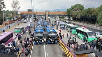Estación de autobuses con varios trolebuses azules, otros autobuses y multitudes de personas. Un puente peatonal elevado y carriles de tráfico son visibles al fondo