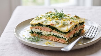 Plato blanco con una porción de lasaña de salmón, ricota y espinaca. La lasaña está cubierta con eneldo, ralladura de limón y un tenedor.