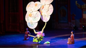 Tres acróbatas en un escenario. Una acróbata manipula seis parasoles decorados con los pies. Dos acróbatas sentadas observan. El fondo tiene decoración de estilo oriental