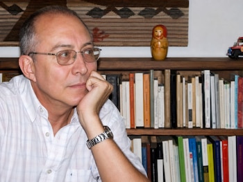 Retrato de Omar Corrado, hombre calvo con gafas y camisa a cuadros, apoyando la mano en su barbilla, frente a estanterías con libros y objetos decorativos