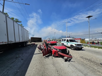 Varios socorristas con uniformes blancos y rojos inspeccionan un automóvil negro destruido junto a la estructura de un camión