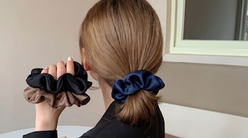 Vista trasera de una persona con cabello castaño recogido en una coleta baja con un scrunchie azul marino, sosteniendo dos scrunchies más (negro y marrón).