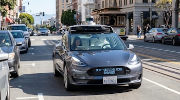 Un coche gris de conducción autónoma con un sensor LIDAR y una pantalla frontal que dice "SELF-DRIVING MODE ENGAGED" se mueve por una calle urbana.