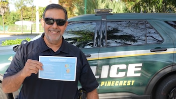 Oficial de policía sonriente con gafas de sol, sosteniendo una tarjeta azul del 'Blue Envelope Program' frente a un SUV policial Land Rover verde oscuro