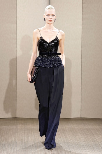 Modelo de tez clara y cabello rubio corto camina en pasarela; usa top con lentejuelas, peplum y tirantes finos, cinturón, pantalones holgados y bolso de mano